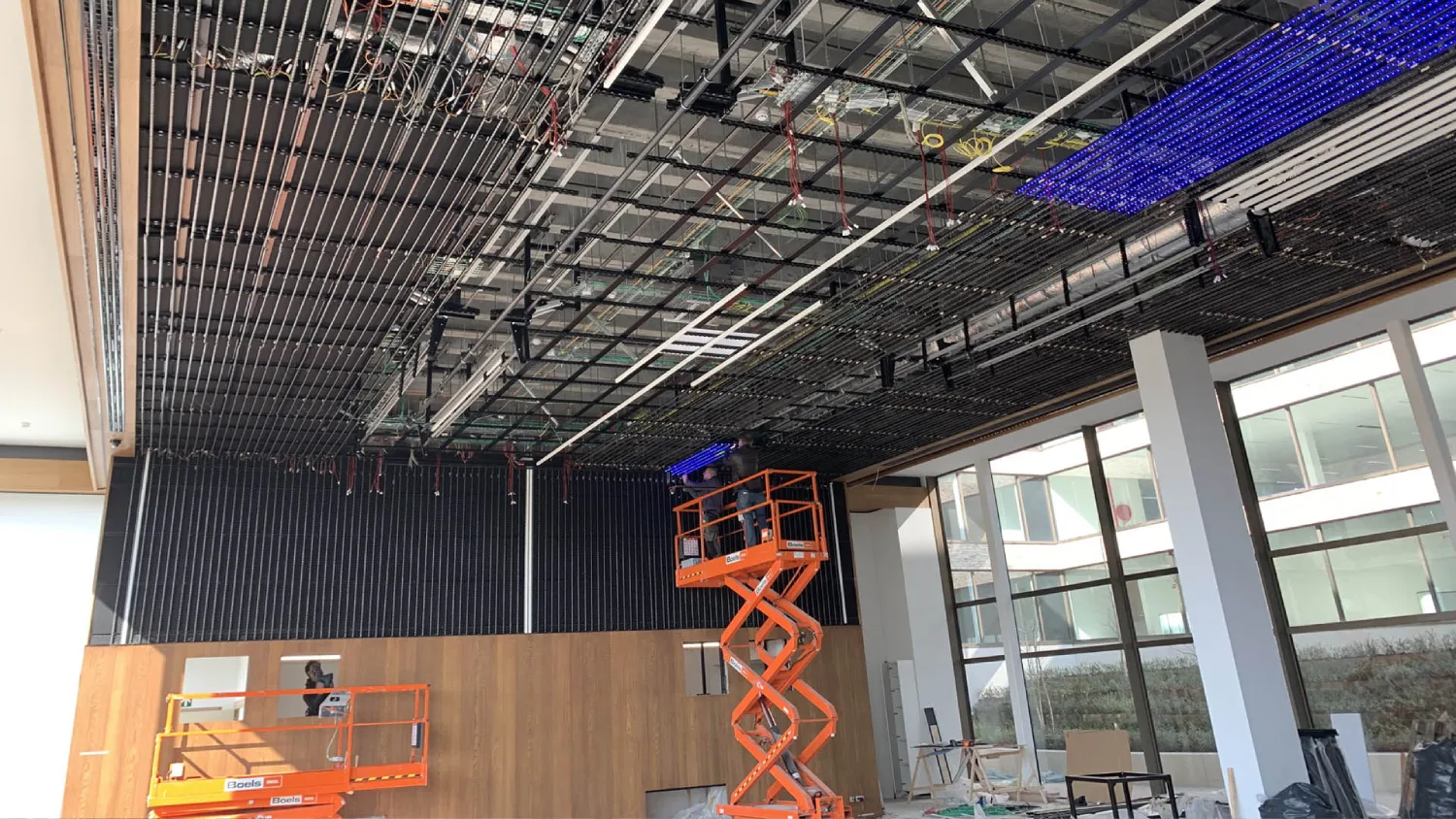Intégration audiovisuelle complète. Vue de face d'une salle avec mur et plafond recouverts d'un réseau dense de barres LED. Au sol, deux nacelles ciseaux orange encadrent des structures de scène (trusses) portant des dizaines de projecteurs lyres asservis abaissés pour maintenance ou installation.